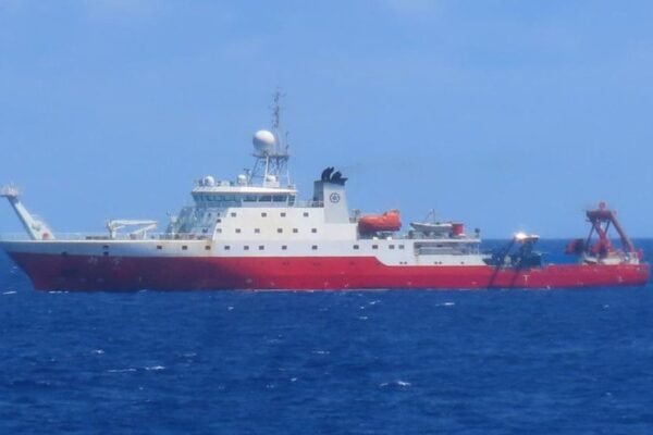 A large red and white research vessel is seen traveling through open ocean, with the horizon line in the background.