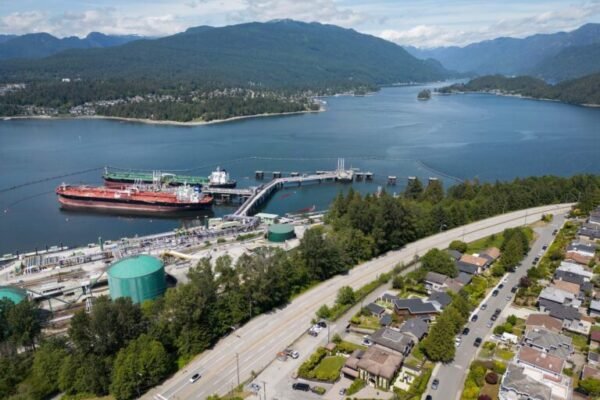 An aerial photo of a petroleum sea terminal, in a landscape of mountains and coastal waterways, with residential homes in the right foreground. There are two tankers docked at the terminal.