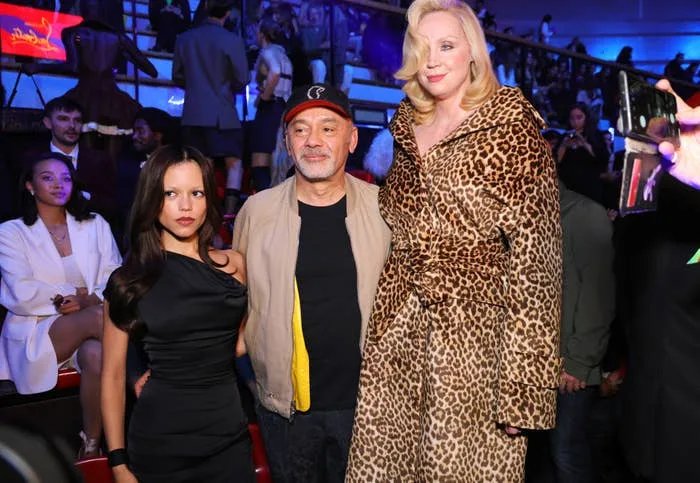 Three individuals pose at an event. A woman in a one-shoulder dress, a man in casual wear, and a tall woman in a leopard-print coat
