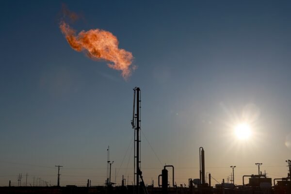 the silhoette of complex factory equiptment lines the bottom of the image, and a tower in the center of the image, also silhoetted, shoots out flames into the sky