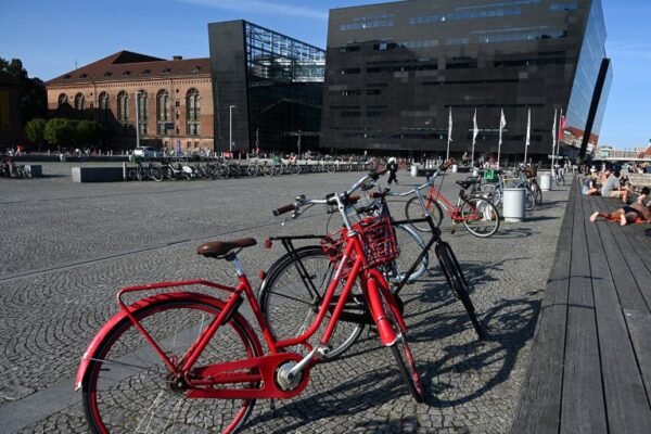 An old-fashioned red bicycle is seen in the foreground of a riverfront promenade in Copenhagen, with the National Library of Denmark in the background.