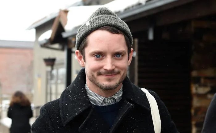 Elijah Wood smiles outdoors wearing a beanie, layered shirt, and coat, with light snow falling around them