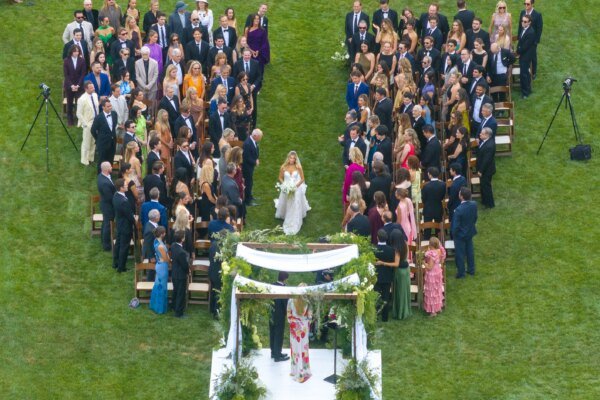 Aerial view of Matthew Lowe and Emily Markus's wedding in Santa Barbara, California, showing guests seated in two curved rows on a lawn, facing a floral-decorated chuppah where the couple stands.