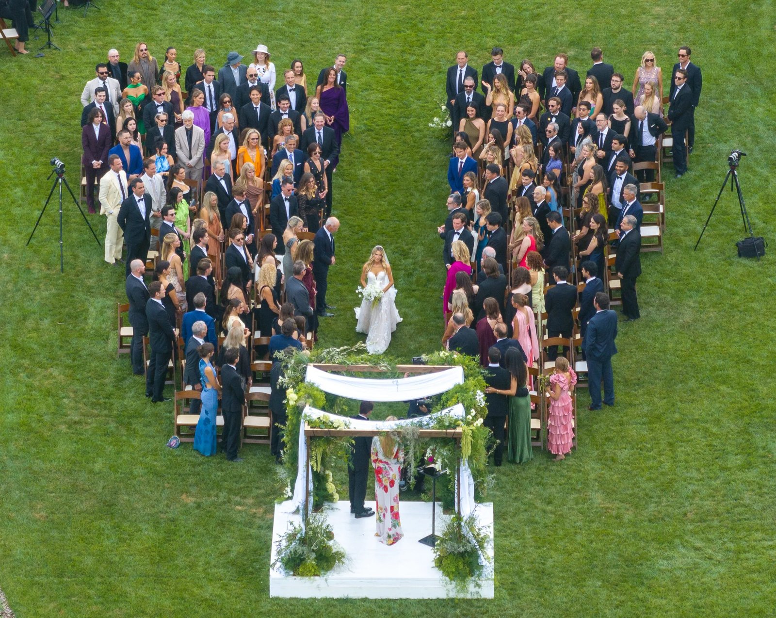 Aerial view of Matthew Lowe and Emily Markus's wedding in Santa Barbara, California, showing guests seated in two curved rows on a lawn, facing a floral-decorated chuppah where the couple stands.