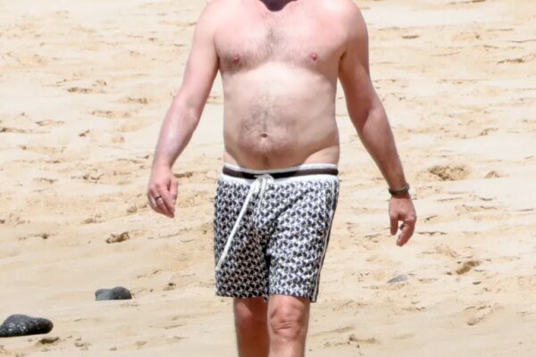 Leonardo DiCaprio walks shirtless along a sandy beach in swim trunks.